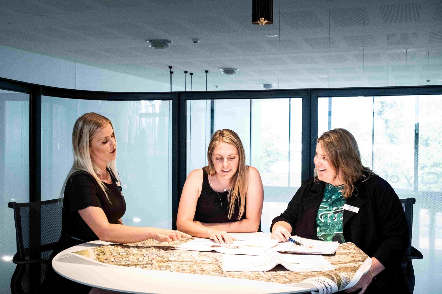 Three ladies, sitting around desk, pointing at maps and smiling