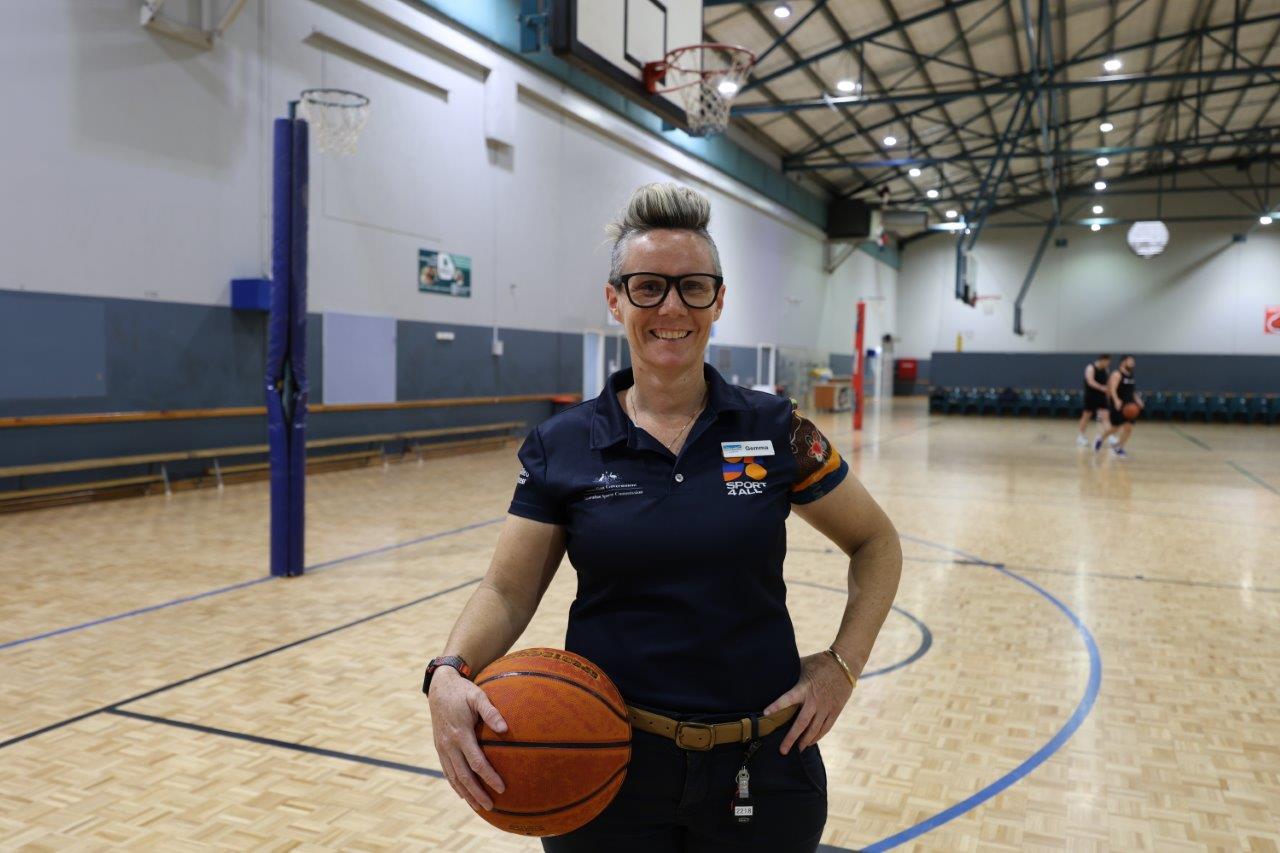 Gemma Harrison, Sports Inclusion Coach stands on the GLC’s indoor basketball court holding a basketball. She wears a navy polo with "Sport4All" logo. In the background, two people are playing basketball.
